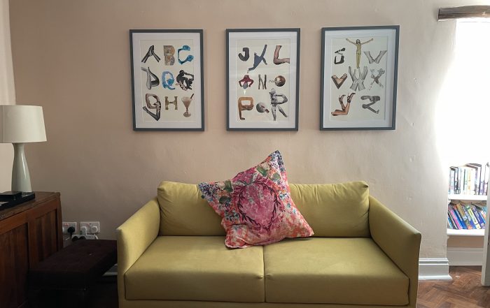 A yellow sofa with a pink cushion. On the wall behind it are framed pictures of collage letters.