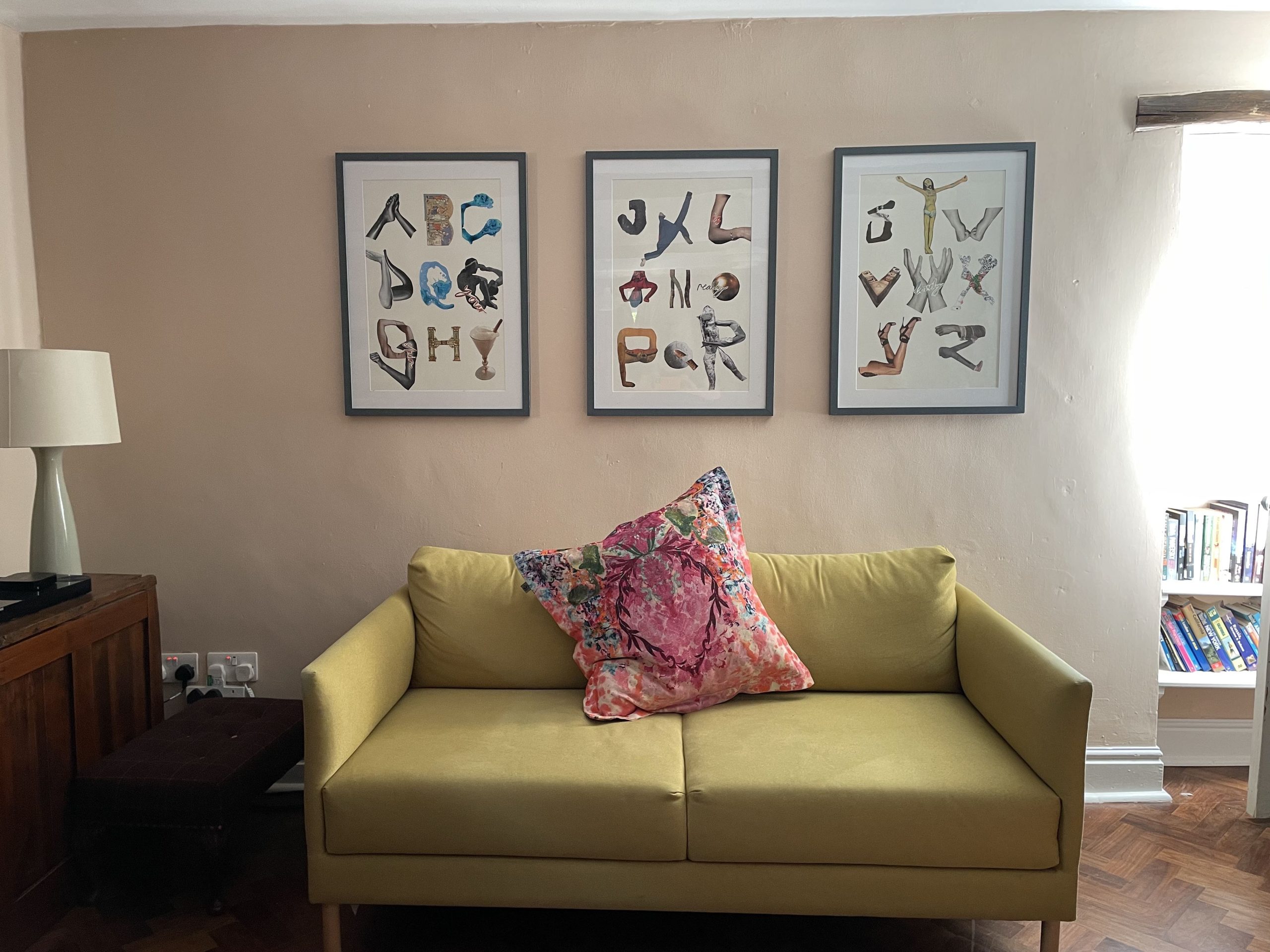 A yellow sofa with a pink cushion. On the wall behind it are framed pictures of collage letters.