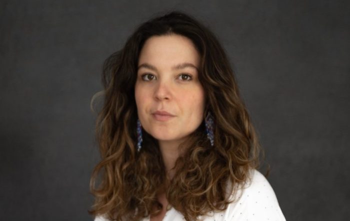 A white woman with shoulder length wavy brown hair and a white shirt.
