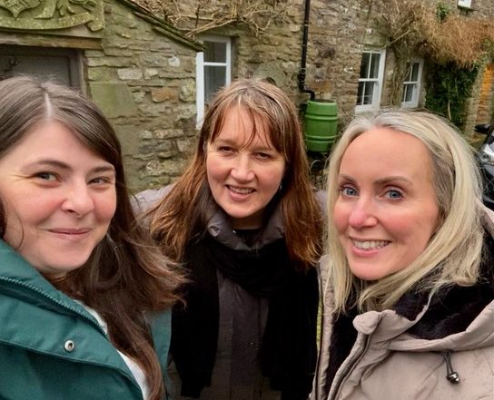 Three white women in raincoats stand smiling against the backdrop of a stone house