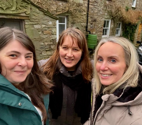 Three white women in raincoats stand smiling against the backdrop of a stone house